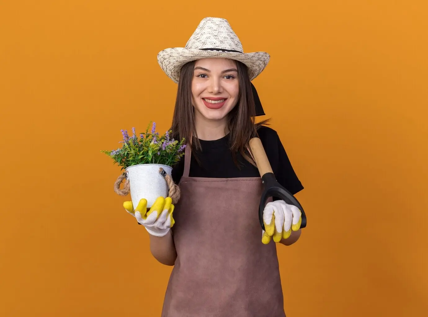 Eine lächelnde, hübsche, kaukasische Gärtnerin mit Gartenhut und Handschuhen hält einen Blumentopf und trägt einen Spaten auf der Schulter vor orangefarbenem Hintergrund.